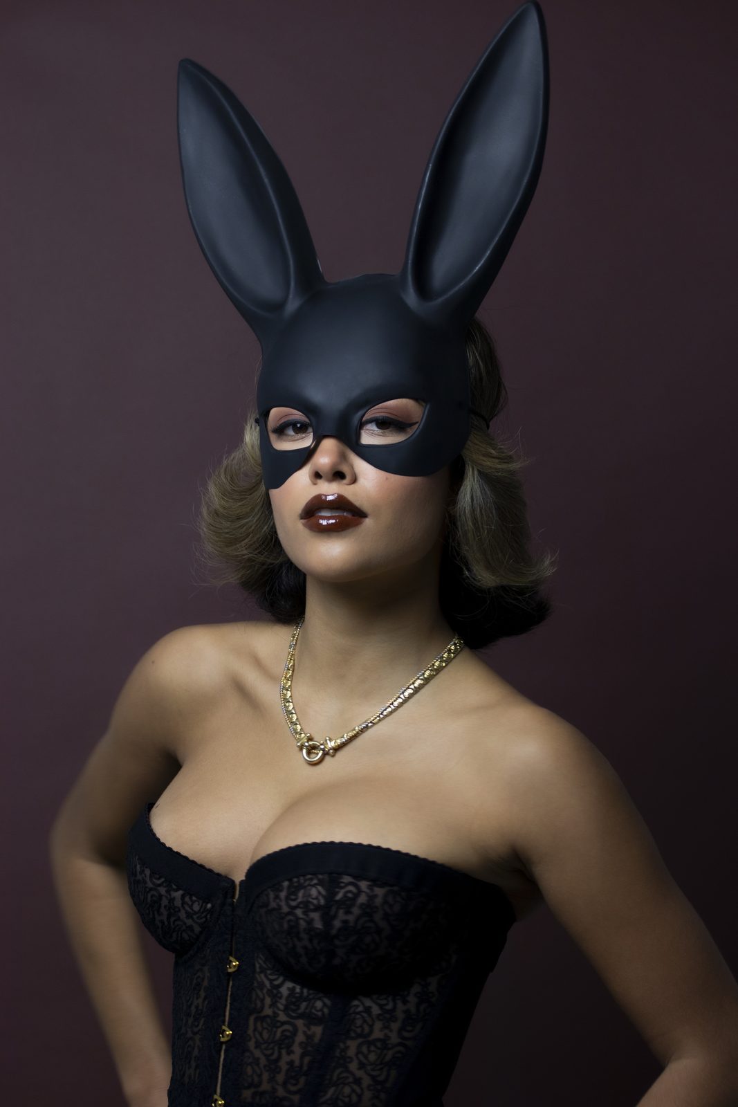 Model in a black bunny mask and lace corset against a burgundy backdrop