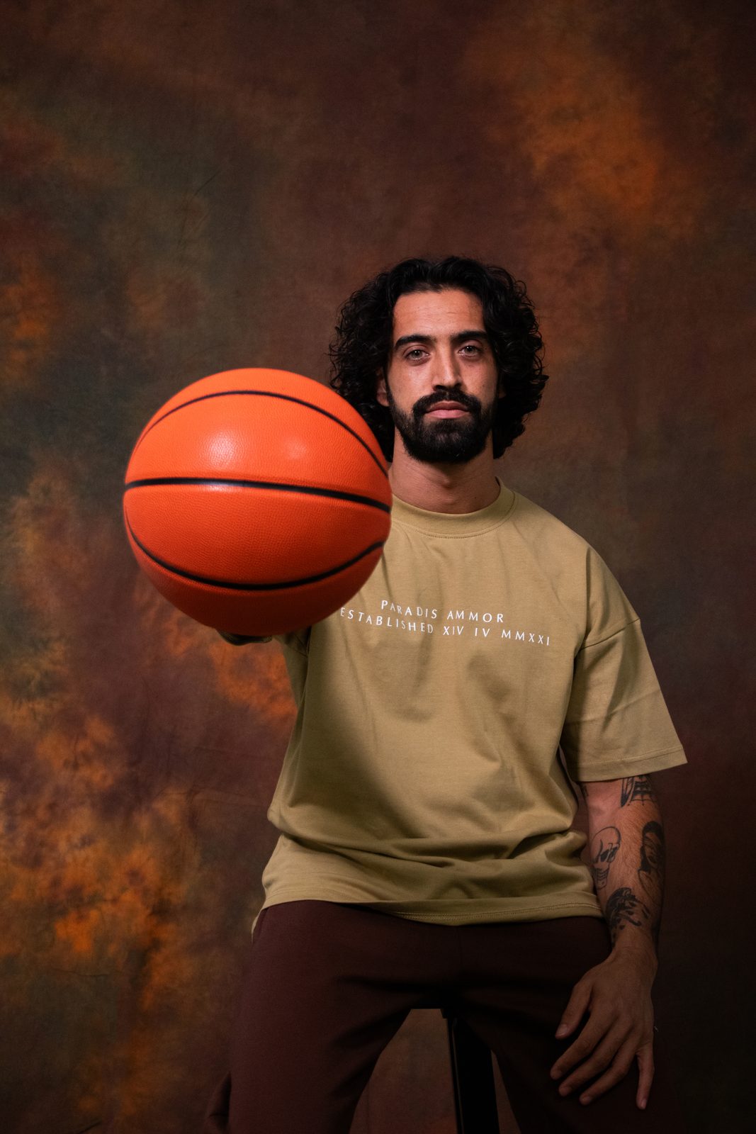 A model in a Paradis Ammor tee and brown trousers tossing a basketball towards the camera in front of a painted rust-coloured backdrop